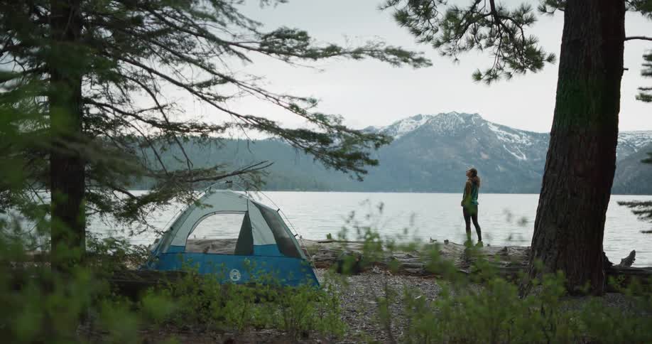 A woman stands next to the shore of a lake in a forest looking around with a camping tent set up behind her at daytime