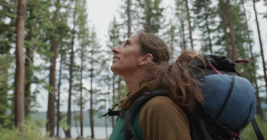 A female hiker looks up at the sky while standing in a woodland next to a lake in the wilderness on a cloudy day