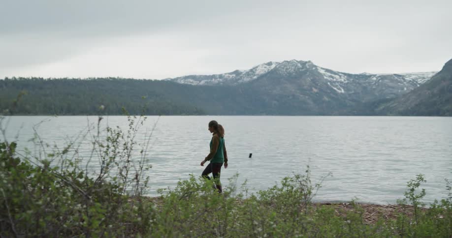 A woman walks along the lake shore looking across at the hills and trees