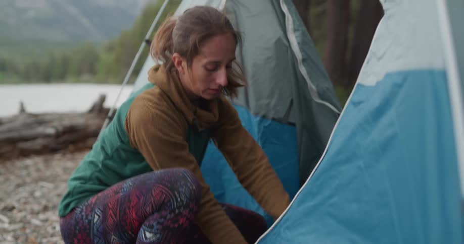 Woman camping near a lake in the mountains opens a tent and takes out a backpack
