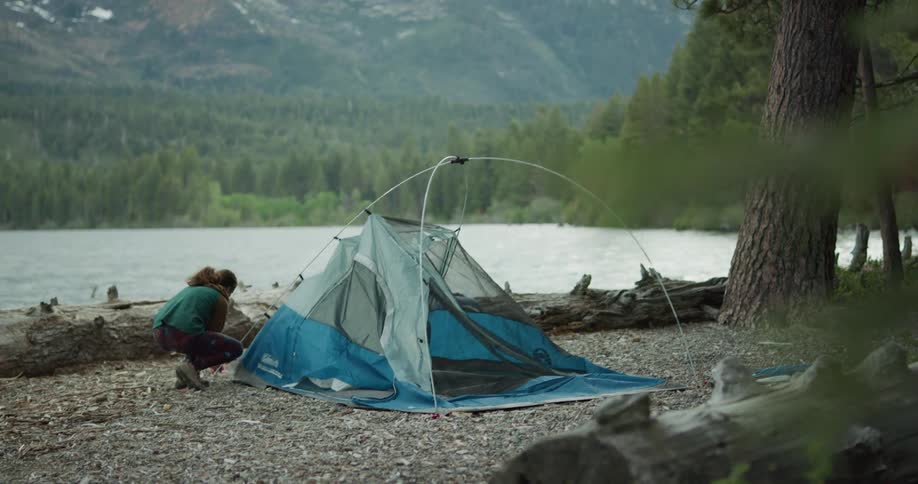 Young woman assembling a tent on the shore of a lake near the pine tree forest in the mountains