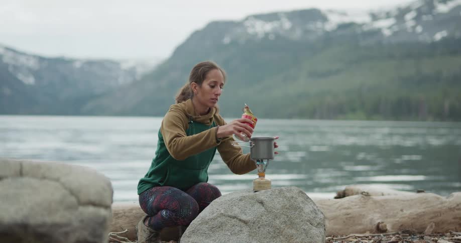 Woman cooking a meal in a small pot over a portable stove while camping near a lake in the mountains