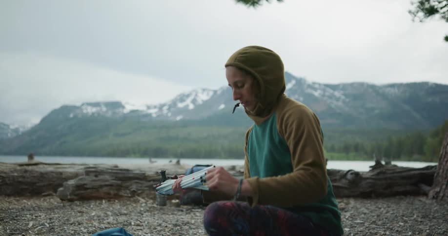Young woman assembling a tent on the shore of a lake near the pine tree forest in the mountains