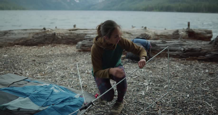 A woman by a lake pitching a tent with a mountain range the sky and clouds in the background
