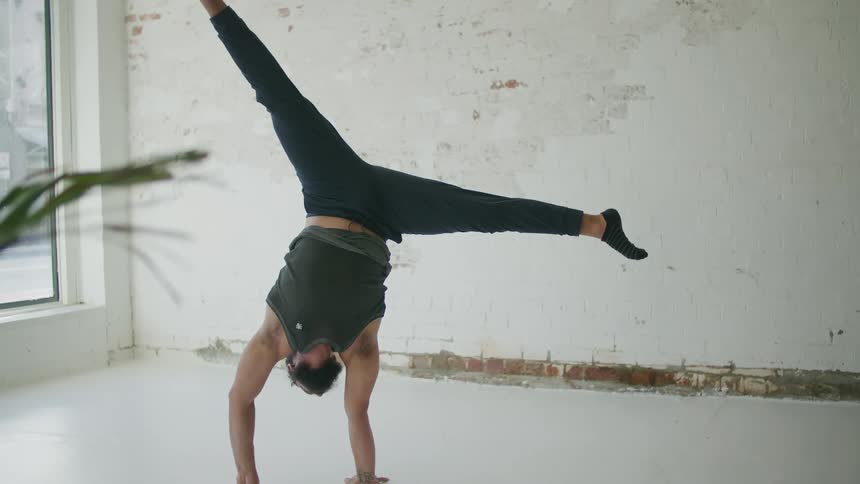Bearded man wearing sports clothes doing yoga stands on his hands in a room