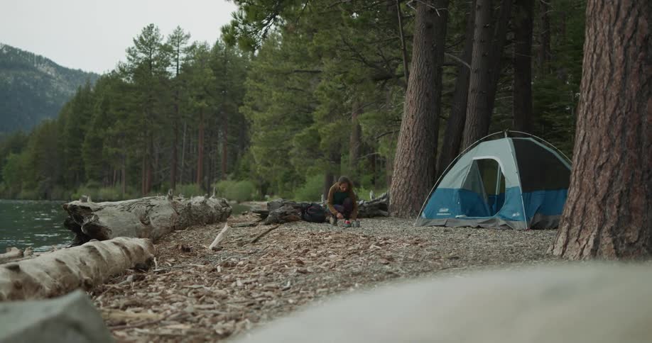 Woman crouches near tent on shore of lake with forest and mountains in background