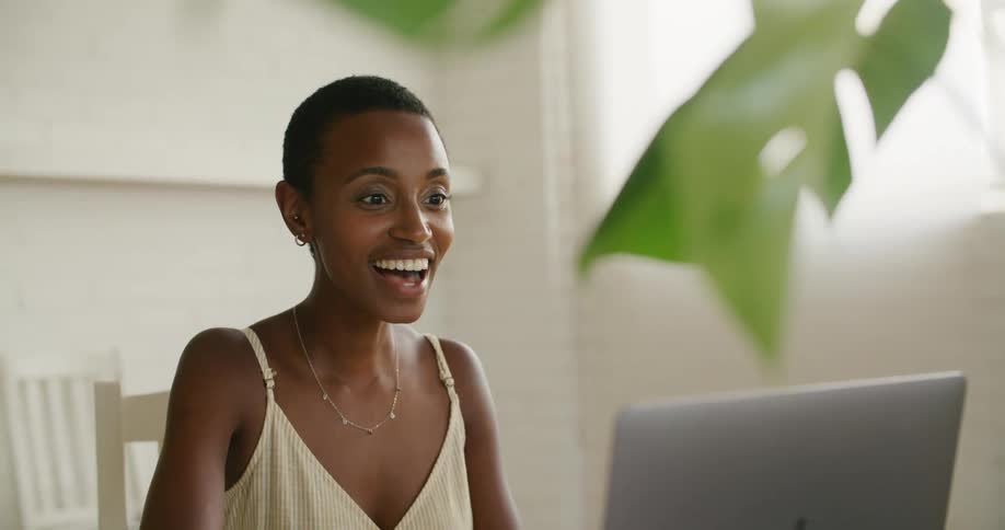 A woman sitting on a chair looking at a laptop with a wall and window in the background