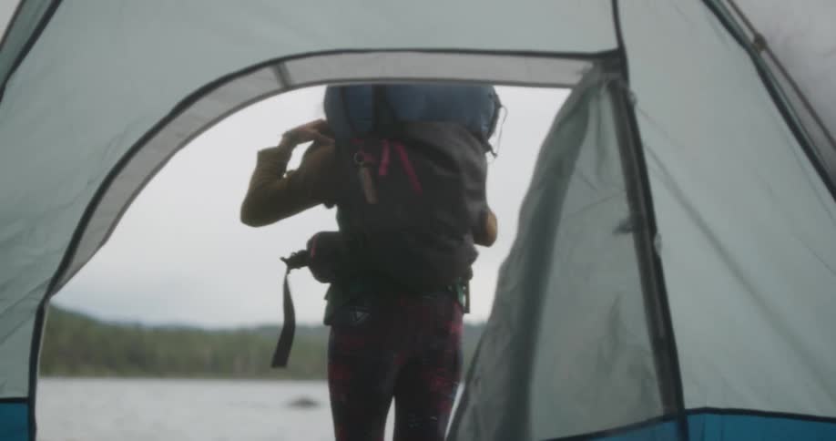 Woman opens tent door to pick up a backpack while camping near a lake in the mountains