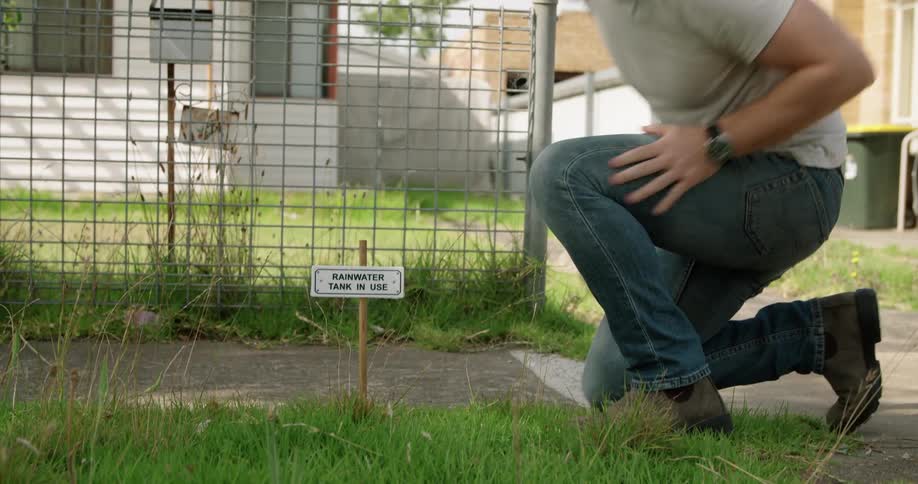 A man uses a mallet to hammer a small sign into the grass outside of the fence around a house during the day