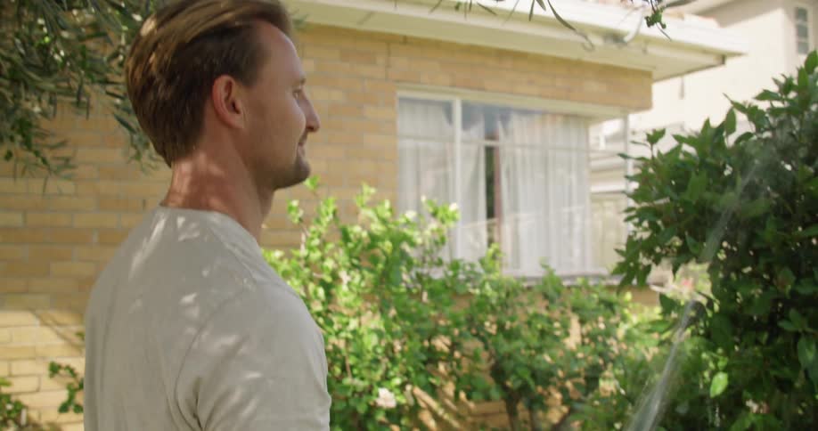Man stands in yard near house watering bush with water hose