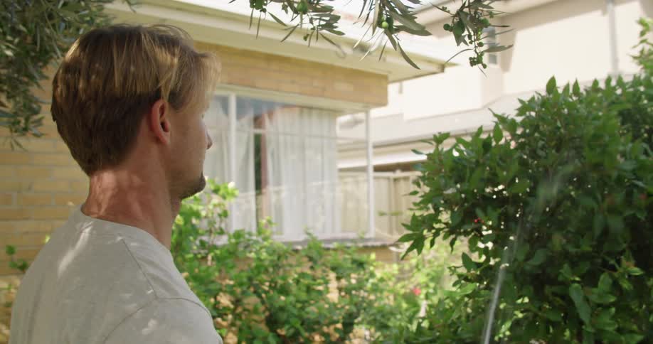 A man uses a water hose to water a small tree in his yard during the day