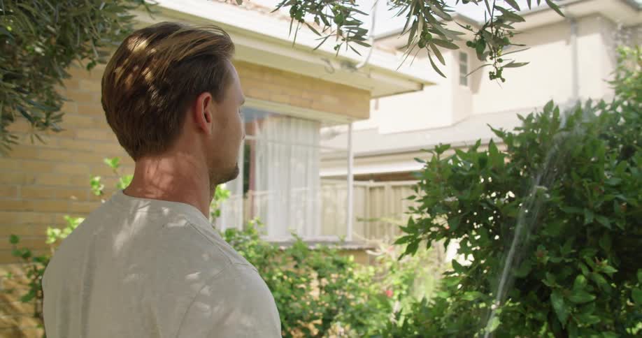 A man stands outside with a garden hose watering a plant
