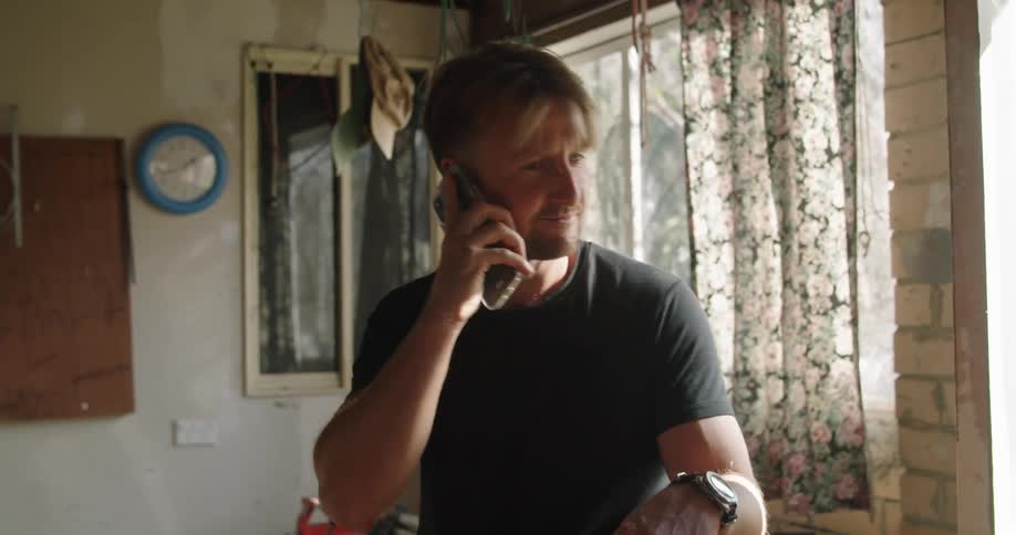 Blonde man with a black t shirt and a wristwatch talks on a smartphone in a room