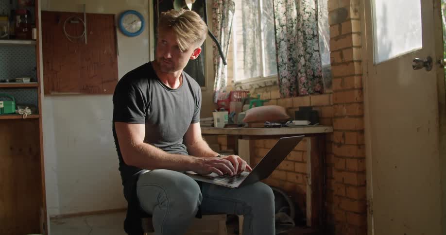 A man in a room sitting on a chair typing on a laptop with a window in the background
