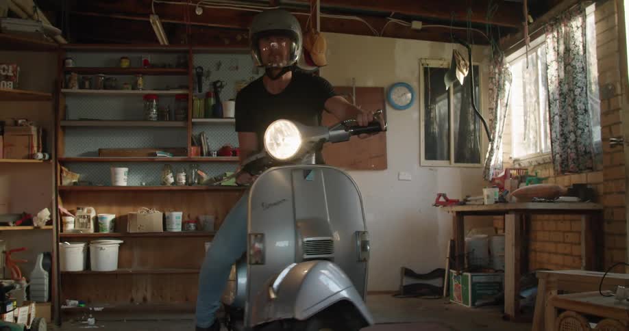 A man wearing a crash helmet drives a vespa scooter out of a garage