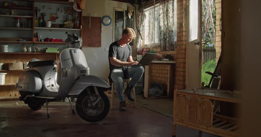 Blonde man with a black t shirt and jeans sits in a chair and uses a laptop next to a white scooter in the garage