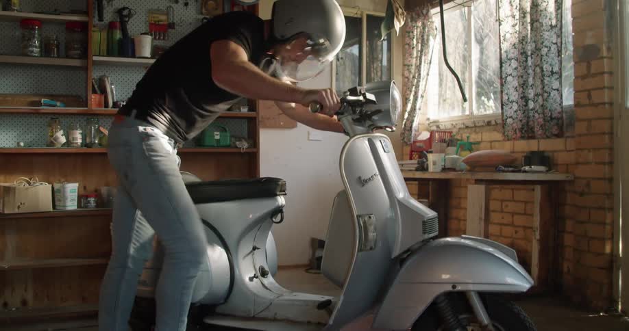 A man in his garage starts his moped and gets on it