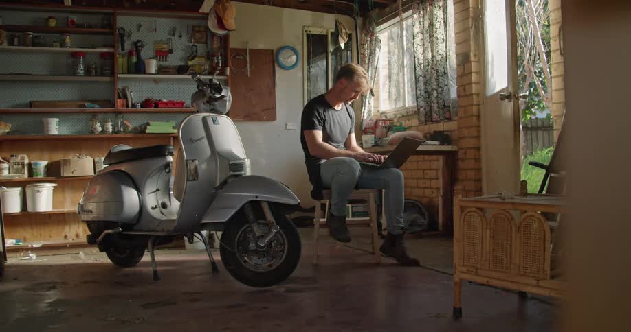 Man sitting in the garage at home close to a vespa scooter typing on a laptop