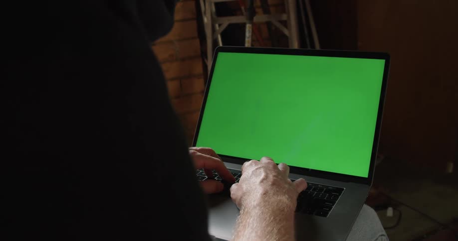 Man sitting inside a dimly lit room works on a laptop with a replaceable green screen
