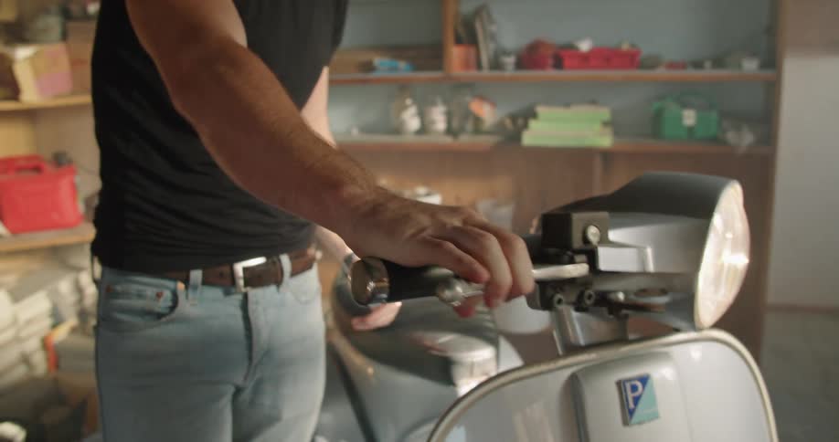 A man in a garage touching a moped with shelves and buckets in the background