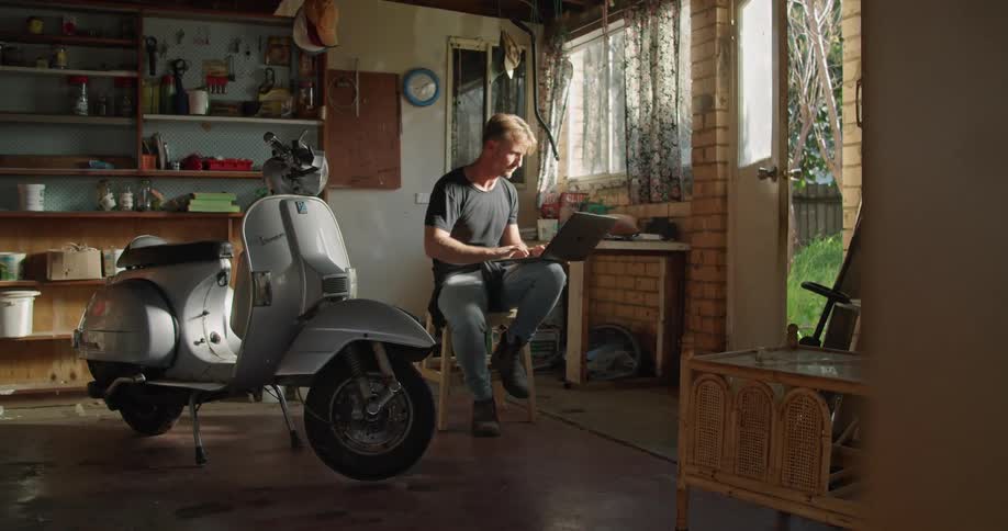 Man sitting in the garage at home close to a vespa scooter typing on a laptop