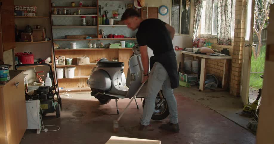 A man sweeps the floor of a garden shed where a moped is parked