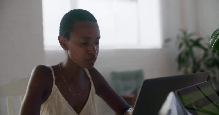 A woman sitting on a chair at a table typing on a laptop with a window in the background