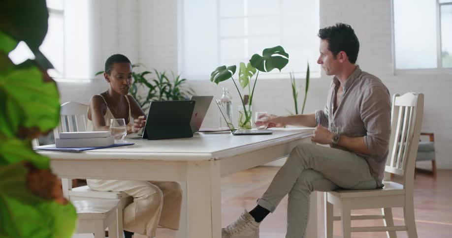 A man sitting in an office passes his computer to a woman across a table and takes a drink of water
