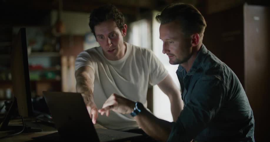 Two men in a room sitting at a table in front of a monitor and a laptop with a wall and door in the background