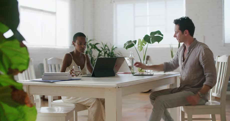 A man and a woman sitting at a table in a room at daytime looking at three different laptops and talking