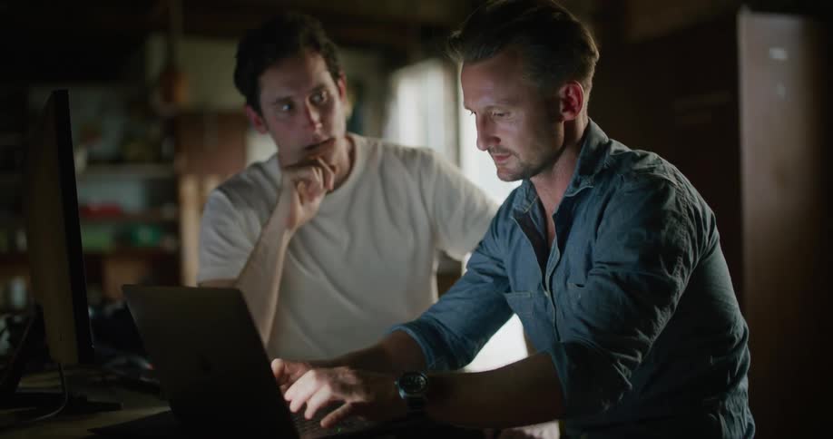 Two men sitting indoors in front of a laptop while one points and the other types