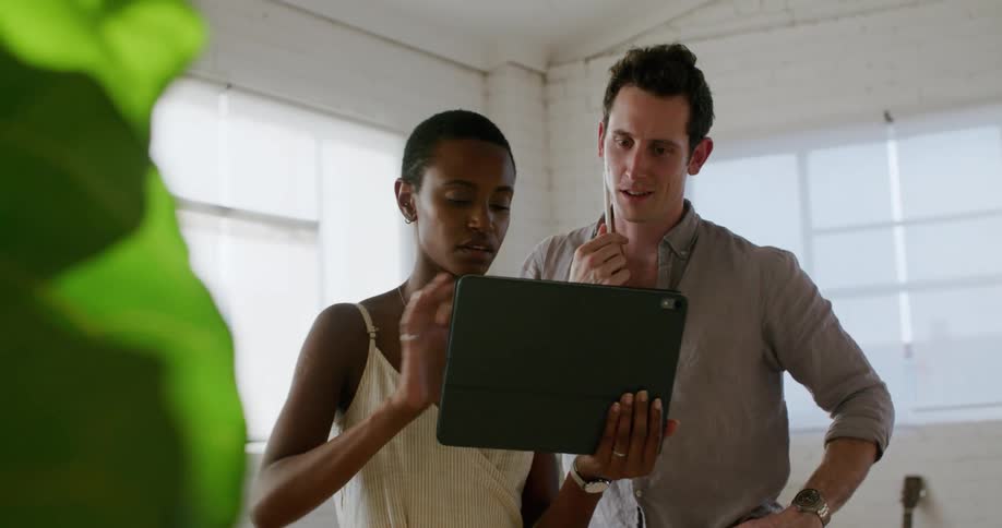 A man and a woman in a room looking at a tablet with a wall and window in the background