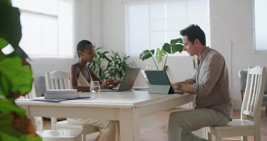 A man and a woman sitting on chairs at a table with laptops giving each other a fist bump with a wall and a window in the background