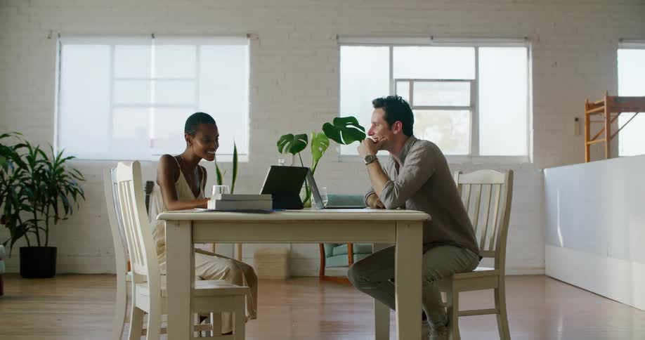 A man and a woman sitting on chairs at a table looking at a tablet and giving each other a high five with a wall and window in the background