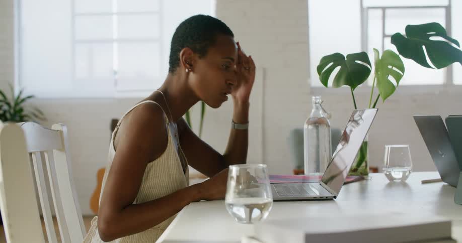 A woman in a room sitting on a chair next to a table typing on a laptop with a wall and window in the background
