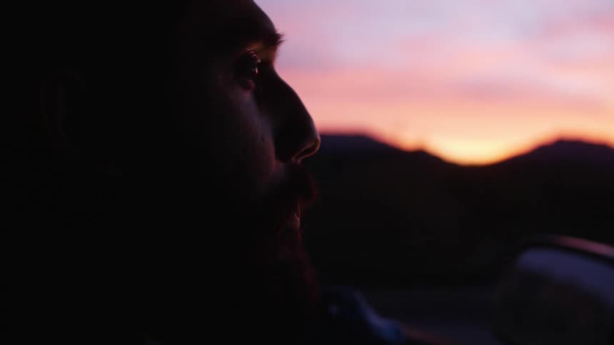 Man with mustache and beard driver vehicle through countryside with red sky and mountains on horizon