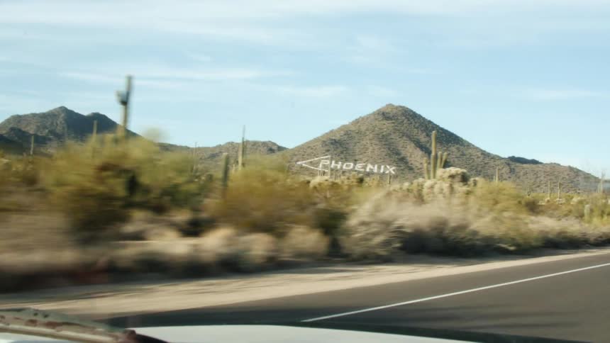 Vehicle drives along road through scrub desert with rocky mountains with the word phoenix on one passing another vehicle