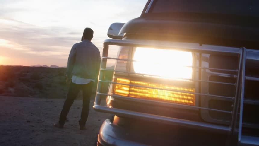 Man looking at the sunset on a rural road behind a car with the headlights on