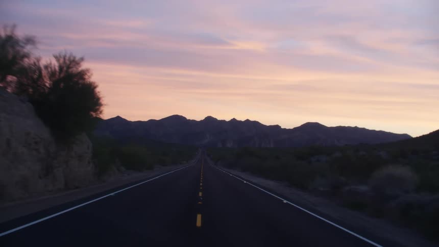 A desert road surrounded by shrubs with a mountain range the sky and clouds in the background