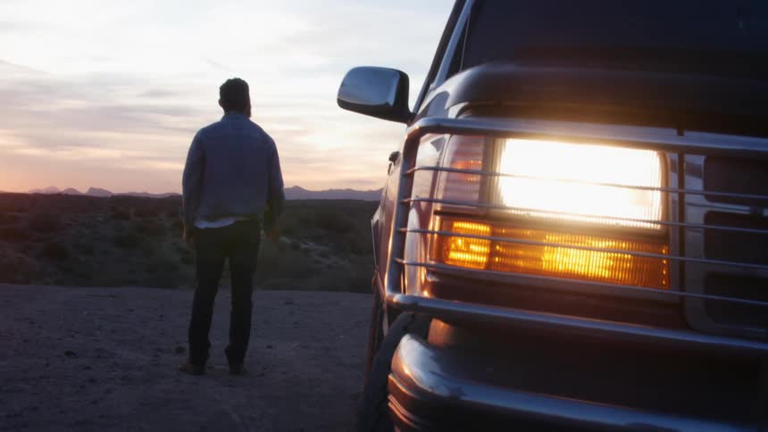 A man standing next to a vehicle with headlights with a mountain range the sky and clouds in the background