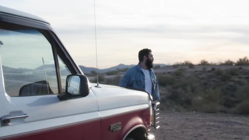 A man takes in the view of the desert and canyon from the hood of his car before hopping down