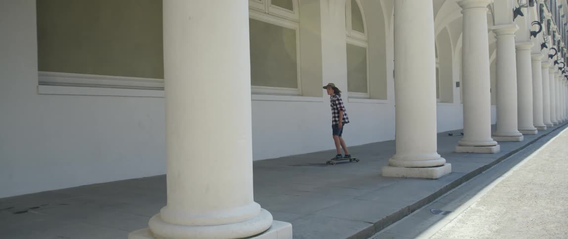 A person with a hat riding a skateboard by a building with pillars