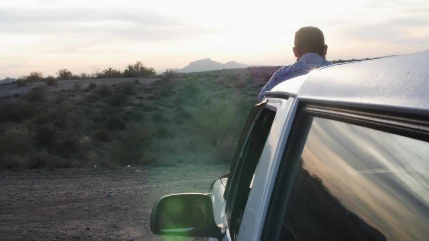 Man looking at the sunset on a rural road reclined on the hood of a car