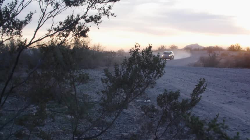 Vehicle with headlights on driving on a dusty dirt road in a rural area at sunset