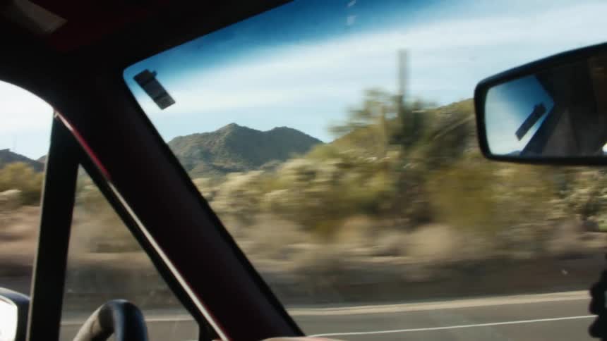 A person in a vehicle with a windshield holding a steering wheel with trees the sky and clouds in the background