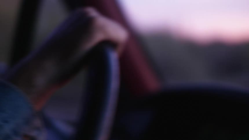 A hand on a steering wheel driving a vehicle at sunset