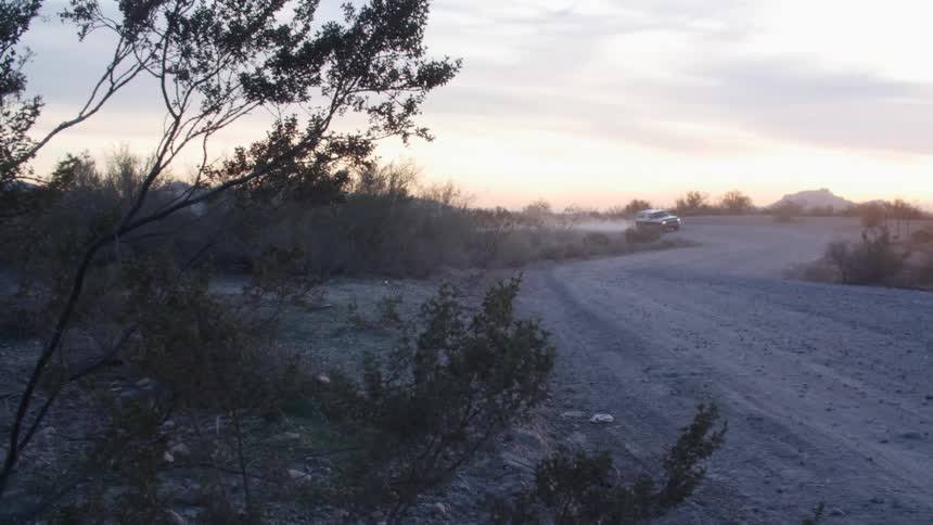 A vehicle driving on a dirt road surrounded by plants and trees with the sky and clouds in the background