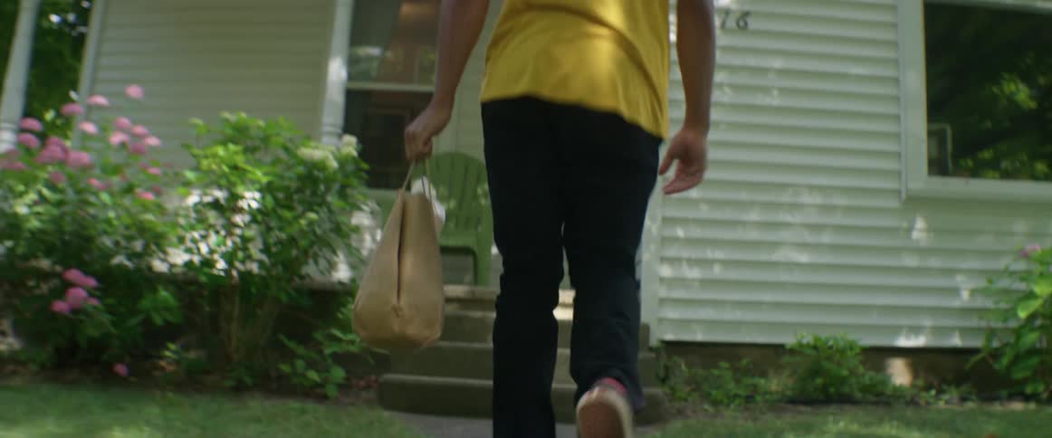 A teenage delivery driver on a bicycle drops off a paper bag of food on the porch of a house