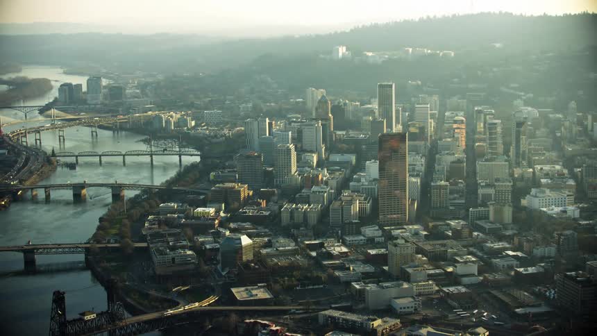 A gritty aerial shot of downtown Portland Oregon showing the Willamette River on the left side of the frame and the downtown skyscrapers on the right side