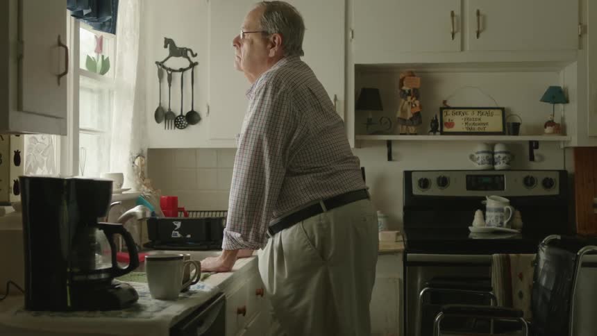 A man standing in a kitchen with a wheelchair behind him with a wall and window in the background